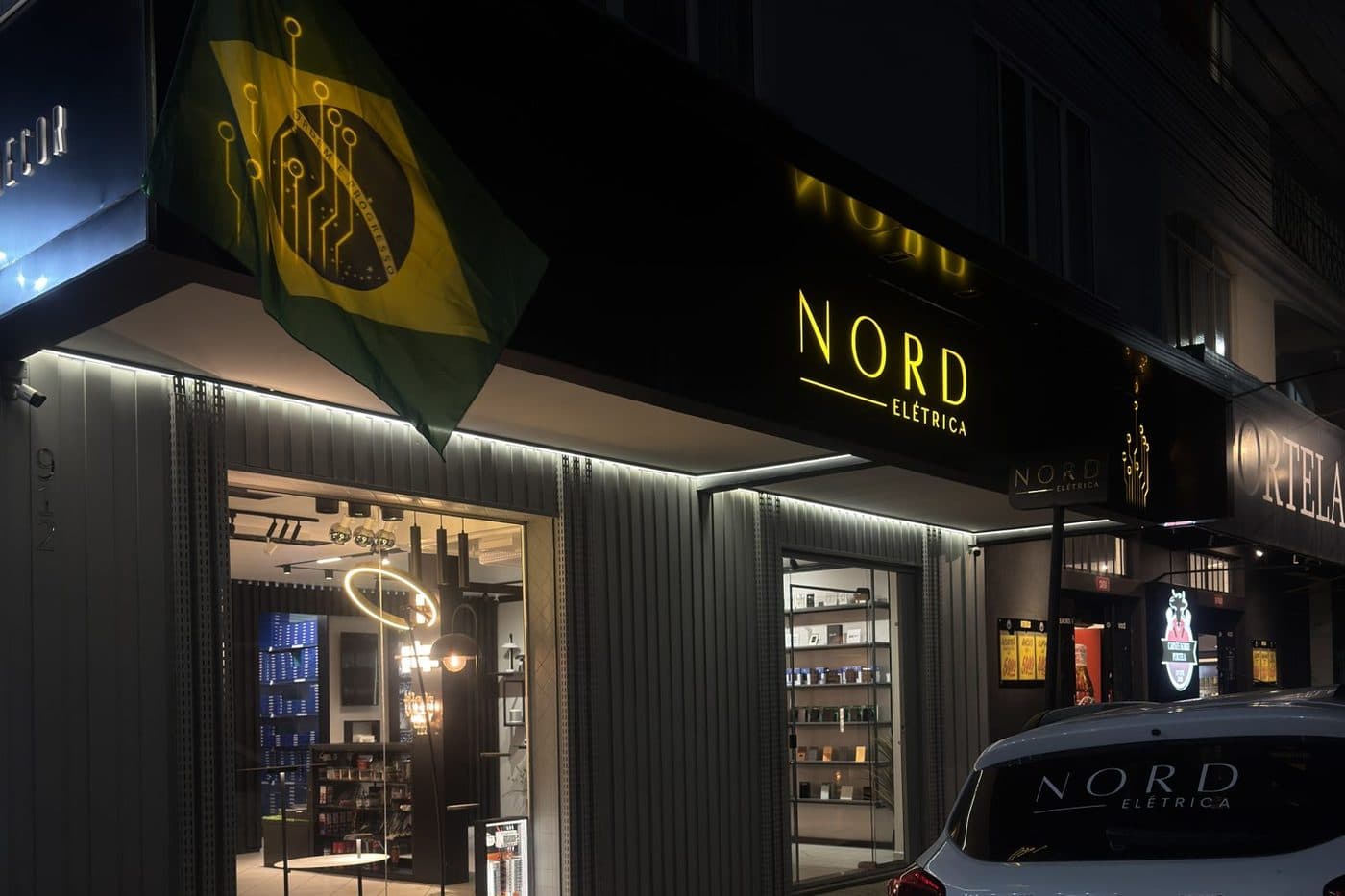 Fachada da loja Elétrica NORD na Rua Sete de Setembro, em Porto União, à noite, com a bandeira do Brasil e o letreiro iluminado